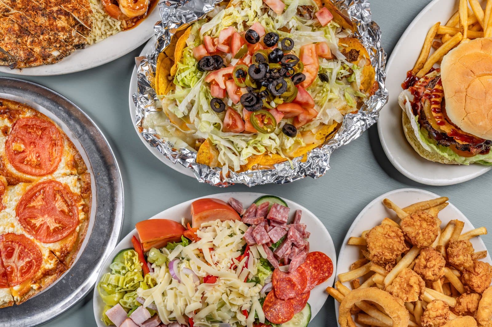 Overhead spread with loaded nachos, salads, pizza, and a burger